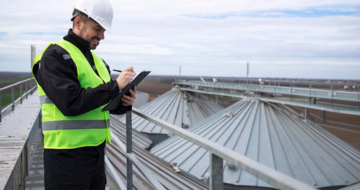 A man in a safety vest stands on a roof, focused on a tablet in his hands, surveying the area below.  