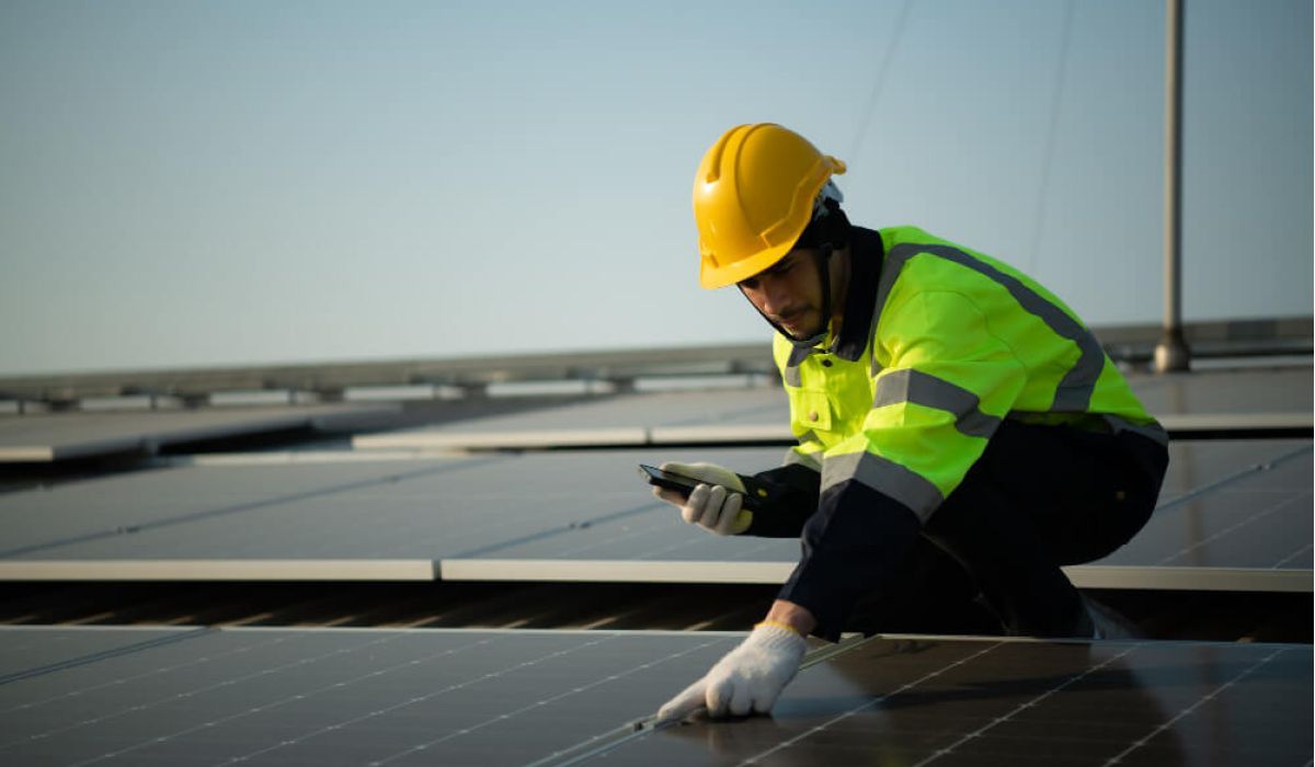 A worker in a yellow hard hat and reflective jacket inspects solar panels on a roof, using a smartphone. The scene conveys focus and modern technology.