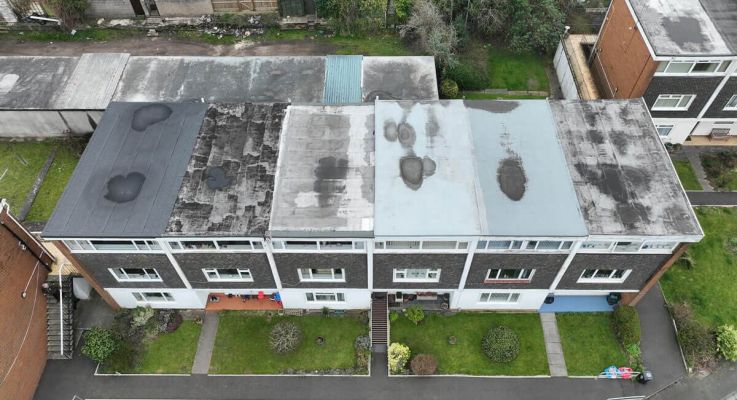 Aerial view of a residential apartment block with multiple flat-roof sections showing weathering, patch repairs, and dark areas, surrounded by lawns, paths, gardens, and neighbouring properties.