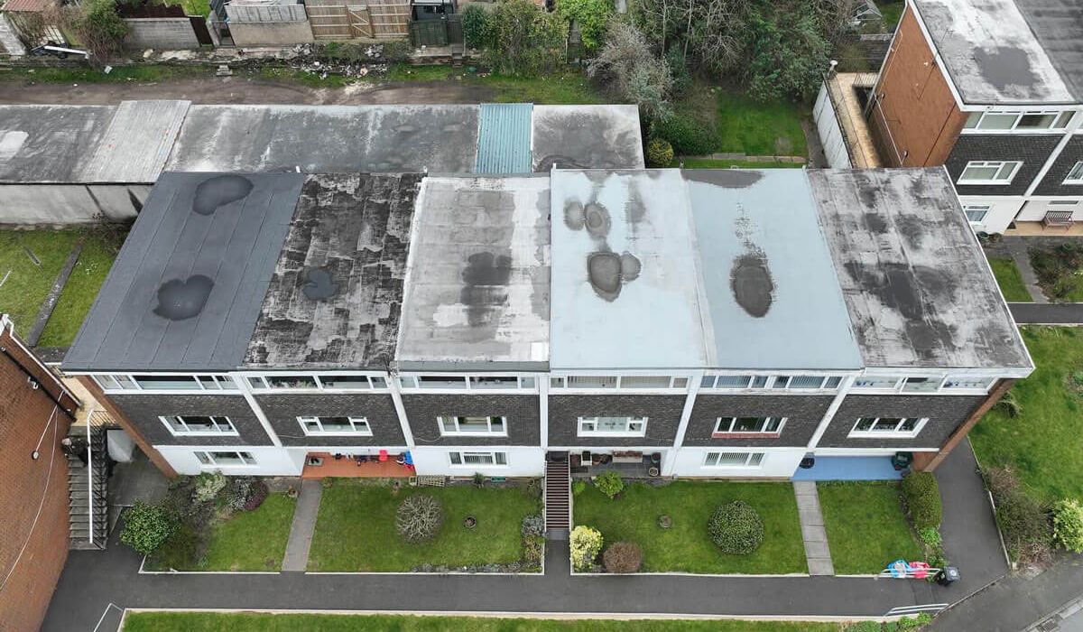 Aerial view of a residential apartment block with multiple flat-roof sections showing weathering, patch repairs, and dark areas, surrounded by lawns, paths, gardens, and neighbouring properties.